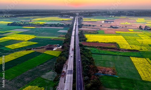 An aerial view of a highway cutting through patchwork farmlands, where green and yellow fields stretch toward the horizon under a soft evening sky in rural India