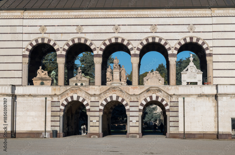 Naklejka premium Striped marble arcade with monumental arches and sculptural tombs at entrance of Monumental Cemetery in Milan, Italy, under clear blue sky.