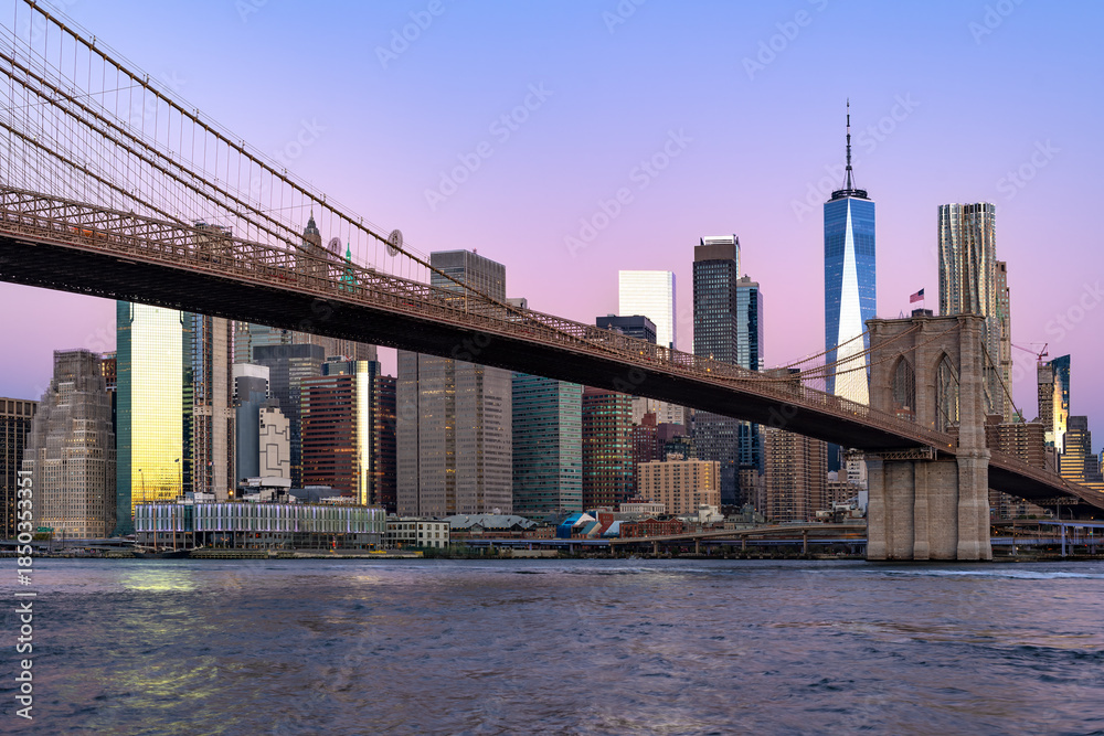 Fototapeta premium The iconic Brooklyn Bridge spans the East River, leading into the Lower Manhattan skyline during a peaceful purple dawn.