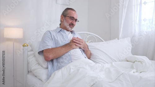 Elderly man sitting on the bed and holding his chest in pain, illustrating symptoms of heart attack, chest pain, or medical emergency at home