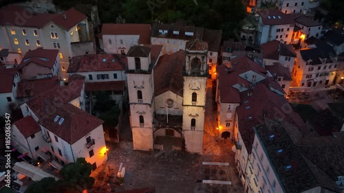 Aerial drone view of the Cathedral of Saint Tryphon surrounded by medieval stone buildings at dusk, showing the historic architecture in warm lighting. Kotor, Montenegro