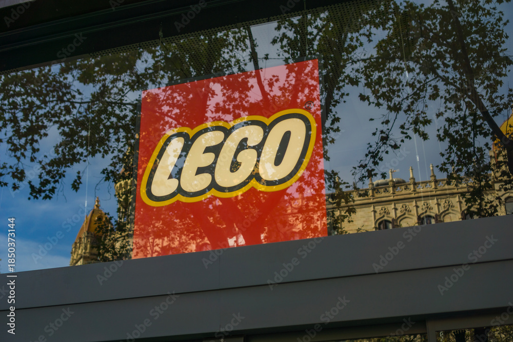 Fototapeta premium Barcelona, Spain, Europe - Nov 23, 2025: lego logo red yellow glass window storefront reflecting trees and historic architecture in a city street under blue sky with copy space for banner design