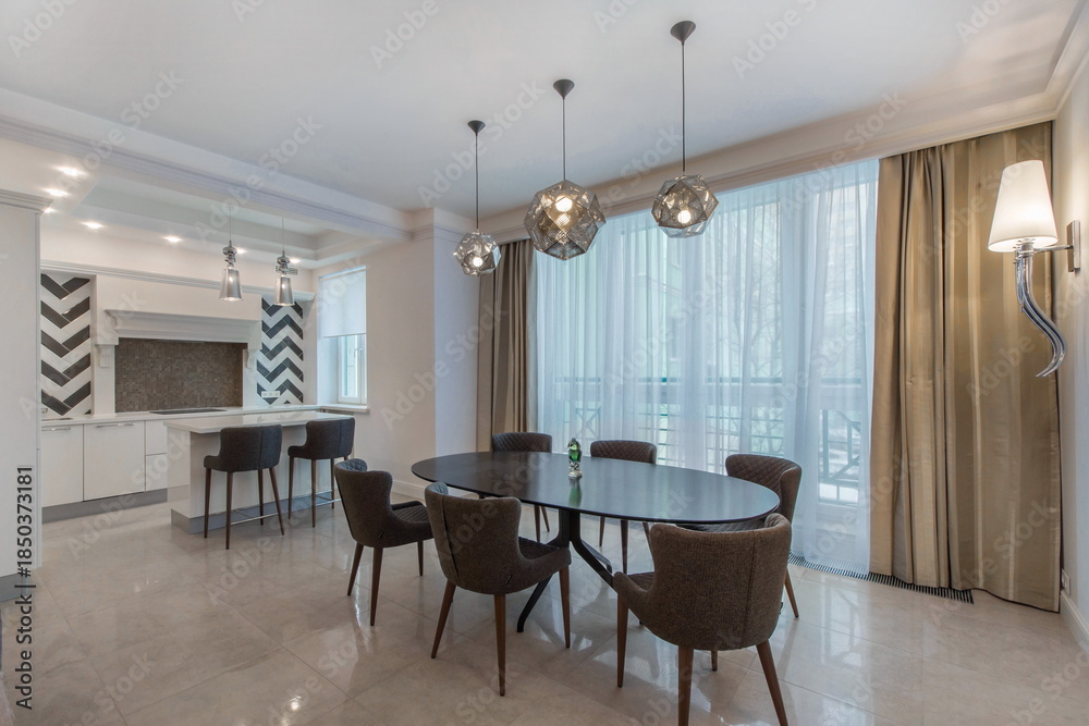 Fototapeta premium A dining area shows a round table with several chairs arranged around it. Natural light comes through large windows. The kitchen is visible in the background with cabinets.