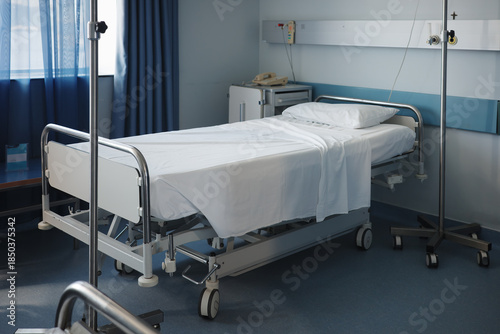 Empty hospital bed with white linens and an intravenous therapy pole standing next to a medical supply cabinet near a blue curtain in a clean clinical ward room ready for the next patient admission