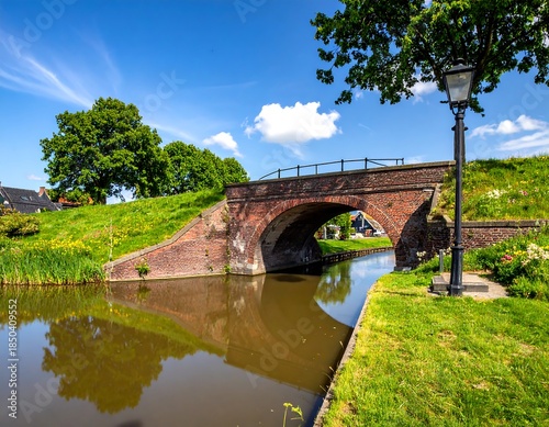 Quaint canal bridge with brick architecture reflecting in the calm water against a backdrop of lush green grass and blue sky