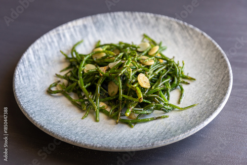 Fried green salty crisp samphire or salicornia plants with garlic on plate on grey background, close up