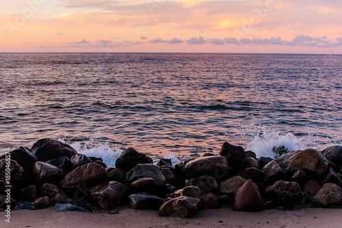 Rocky shoreline with gentle waves at sunset, pastel sky reflecting on the sea. Calm coastal landscape with stones, water, and soft evening light.