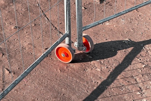 Orange wheels of a temporary metal fence casting long shadows on dusty ground, highlighting construction textures and urban infrastructure.