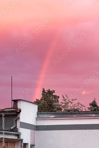 Soft pink evening sky with a faint rainbow rising above rooftops and trees, creating a dreamy and atmospheric urban landscape.