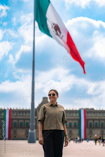 Smiling young Latin woman standing at Zocalo of Mexico City with the huge Mexican flag waving in the background under a bright blue sky, wearing casual clothes, sunglasses, showing national pride