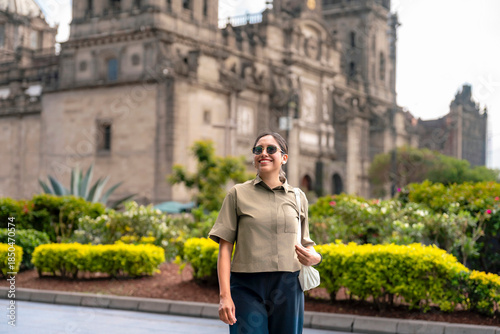 Smiling young Latin woman walking relaxed near the Metropolitan Cathedral in Mexico City’s Zocalo, surrounded by green gardens and bright daylight, wearing modern clothes, sunglasses under a blue sky