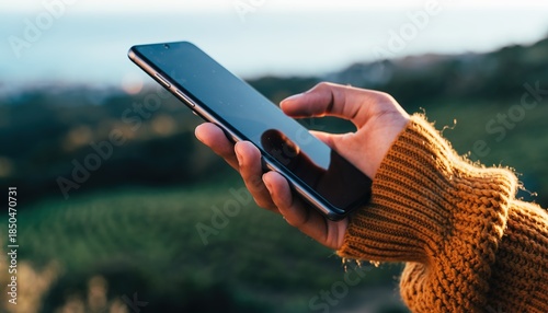 Close-up of a persons hand holding a smartphone outdoors with a scenic landscape in the background.