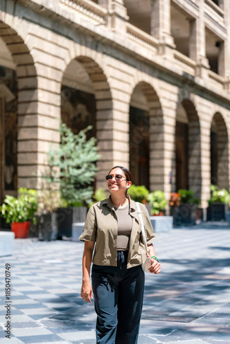 Smiling young Latin woman walking at courtyard of Museo Vivo del Muralismo in Mexico City, enjoying the sunlight and historic architecture, surrounded by plants, stone arches, and artistic atmosphere