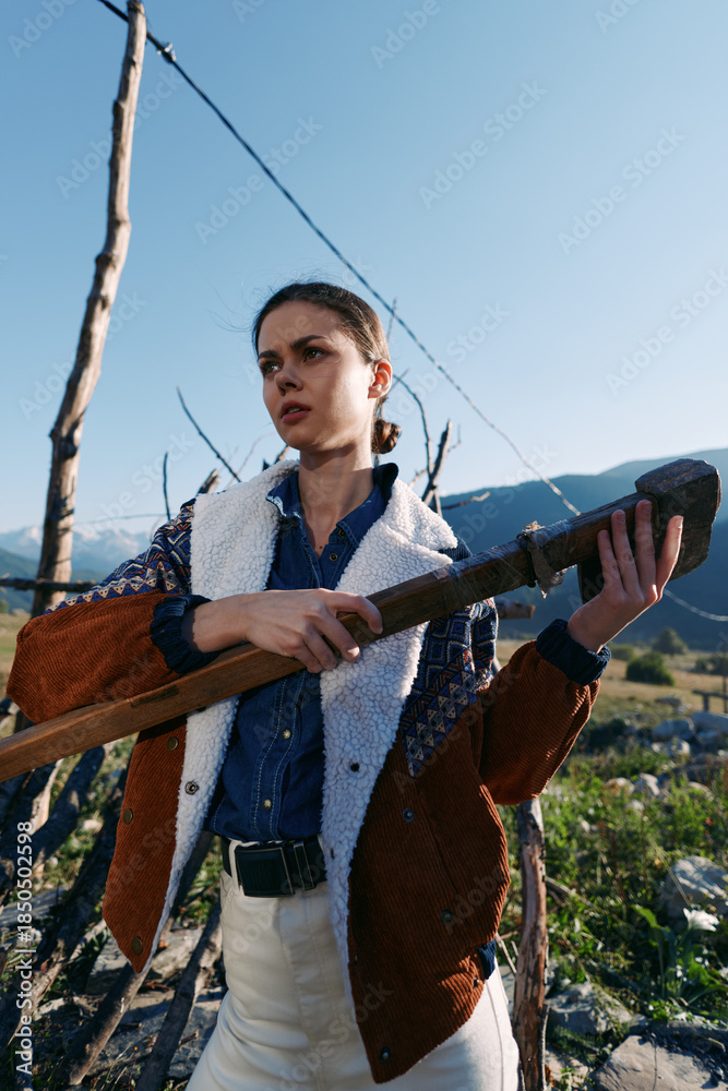 Fototapeta premium Woman holding a pole outdoors in mountains, rural portrait of a young female worker in jacket and denim, standing in a field with determined expression under clear sky and sunlight