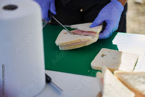 Foto Gloved Hands Slice Ham Sandwich On Green Cutting Board Outdoors