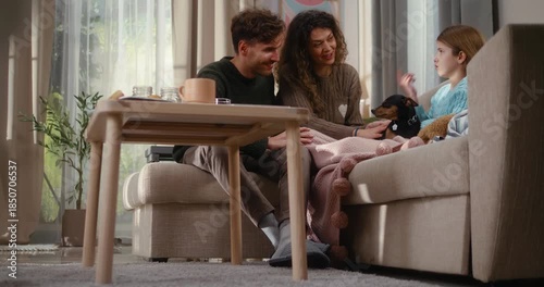 Happy parents sit together on a cozy sofa, talking with their daughter who is holding a dog. Warm family moment, comfort, love, and everyday life in a modern living room.