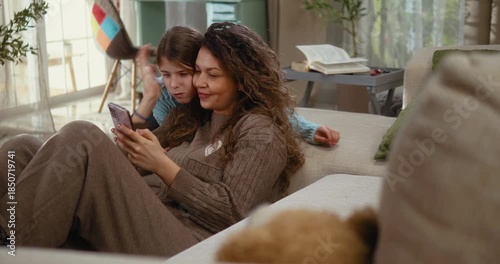 Mother and daughter sit comfortably in the living room looking at a smartphone screen and chatting together. Relaxed family moment, connection, laughter, and free time at home.