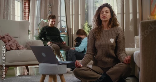 Mother working online from home takes a mindful break and meditates on the living room floor. In the background, father and daughter play with the dog, showing balance between work, family, and wellbe