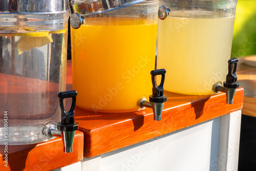 Three large glass beverage dispensers with chrome lids and taps on a wooden counter, filled with water, orange juice and lemonade, ready for self service at a cafe or event