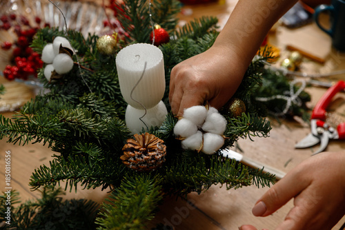 Tableau sur toile Close-up of woman's hands decorating a handmade Christmas floral arrangement with white cotton flowers, white candle, and pine cones in a cozy workshop