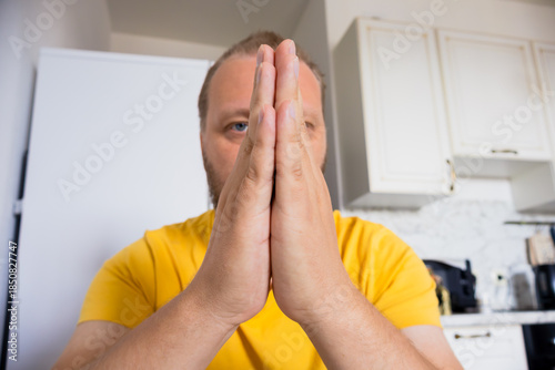 Canvastavla Adult man in a yellow shirt holds his hands together in a prayer or pleading gesture while looking at the camera in a home kitchen