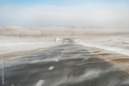 Leere Landstraße in der winterlichen Steppe der Mongolei aus Fahrersicht mit Schneeverwehungen und einer Siedlung und Bergen am Horizont