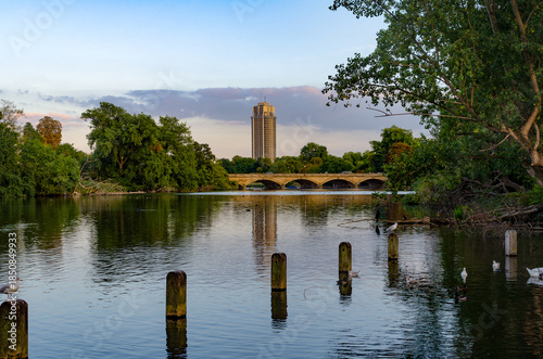 View of a city building over a calm river with trees and a bridge in the background during early evening hours .