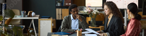 Multiethnic group of professionals discussing business documents at office table, Black man listening attentively while Caucasian woman explaining project details to another woman, header