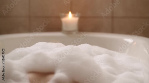 Pov from a bathtub looking along the body covered in white bubbly foam towards a candle burning on the edge with steam rising against a blurred tile wall, concept of a relaxing bath and self-care