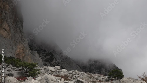 Cloud mountain fog obscures a dramatic sheer cliff and rocky scree slope on a moody, eerie day.