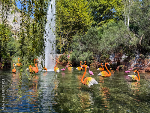 Vibrant Display of Orange and Pink Flamingo Statues in a Lush Green Pond with a Tall Water Fountain and Overhanging Willow Branches