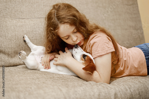 Happy girl and cute puppy dog, kissing and embracing with love and care on couch in lounge at home.