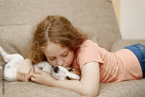 Happy girl and cute puppy dog, kissing and embracing with love and care on couch in lounge at home.