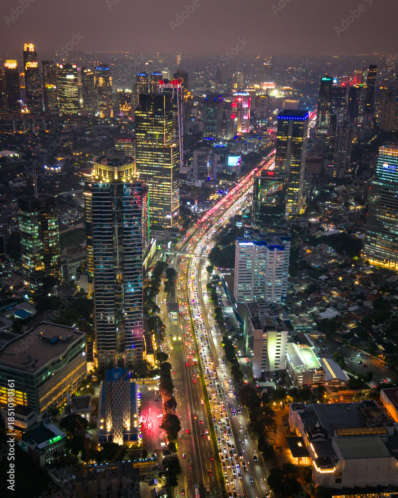 Fototapeta premium Drone aerial view of a vibrant capital city at night featuring illuminated skyscrapers and busy traffic below. Urban cityscape with glowing lights, modern architecture, and dynamic nighttime atmospher