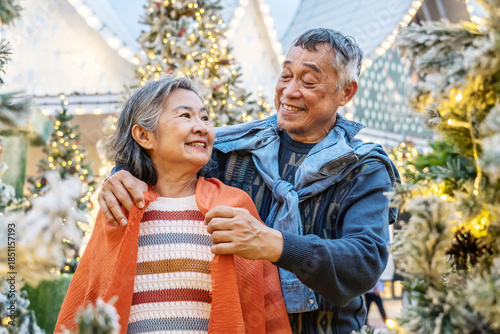 Elderly asian couple embraces near a beautifully decorated Christmas tree. The husband places a warm scarf around his wife, filling the air with love and festive cheer as they share joyful smiles.