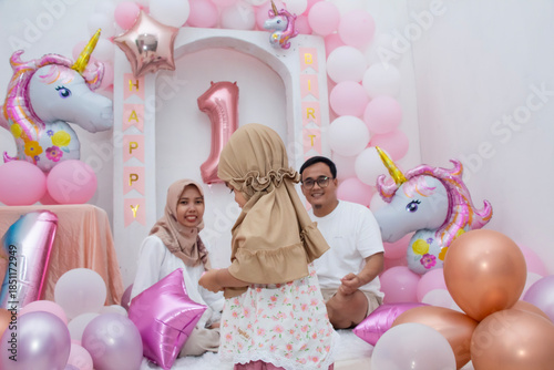 A one-year-old girl wearing a hijab is seen from the side walking without looking at her parents near her birthday decorations.