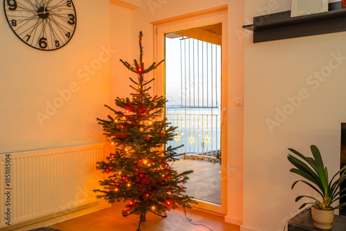 A Christmas tree with red and yellow lights placed indoors near balcony door overlooking the sea at sunset