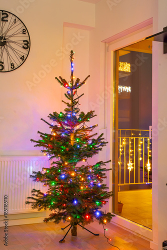 A natural Christmas tree decorated with colorful LED lights stands in a modern living room near a balcony door