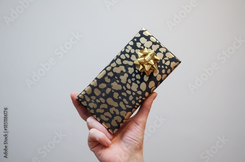 Hand holding a gift box on white background. Black and gold wrapped gift. Shiny gold gift bow.
