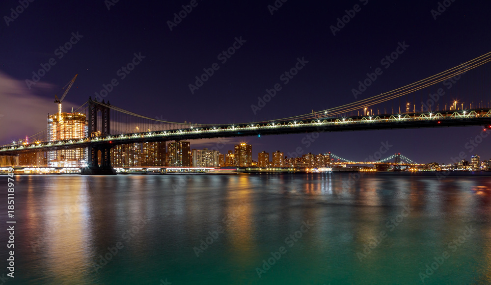 Obraz premium Over East River as Manhattan Bridge lights illuminate night, reflecting on water with New York buildings in distance.
