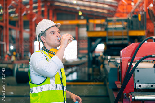 Thirsty Caucasian industrial engineer man wear safety helmet vest drinking water from plastic bottle in factory. Tired worker taking break refreshment. Manufacturing plant hydration health care