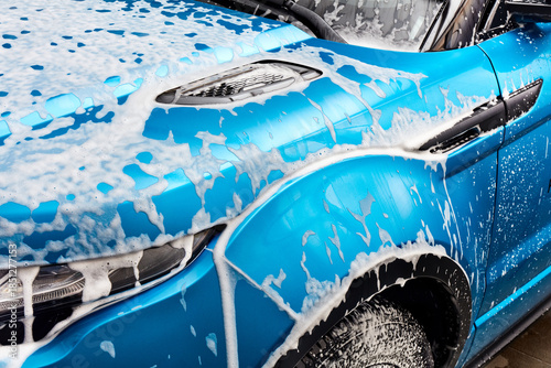 A bright turquoise car is being washed. Thick white foam covers the hood, headlight, fender, and tire. Soap is dripping down the side of the clean automobile.
