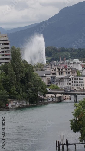 Geneva city view featuring the Jet d'Eau, urban architecture,  bridge over Rhone river and Alpine mountains on a cloudy day