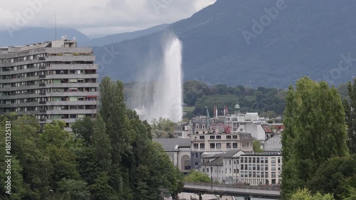 Geneva city view featuring the Jet d'Eau, urban architecture,   and Alpine mountains on a cloudy day