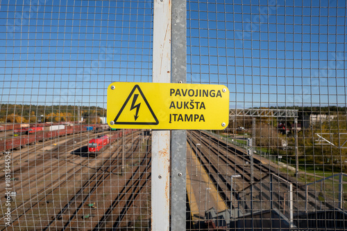 Yellow High Voltage Warning Sign on Safety Fence at Paneriai Railway Yard in Vilnius, Lithuania