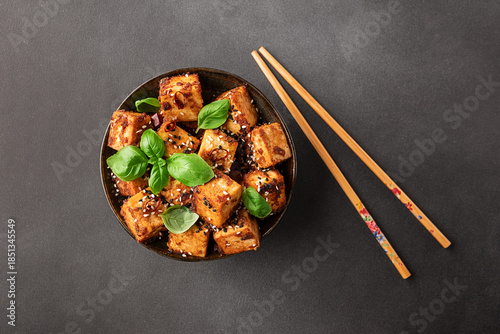 Fried tofu with sesame seeds, soy sauce, spices decorated with fresh basil leaves on black stone background top view. Healthy Asian food concept. Vegan food.