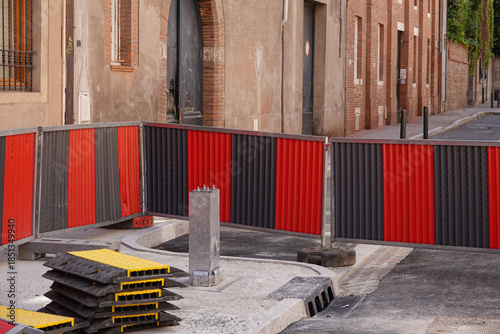 Fototapeta Naklejka Na Ścianę i Meble -  Construction barriers in red and gray surround a work area on a street in Toulouse. The surroundings feature brick buildings and a clear sky, indicating ongoing urban development