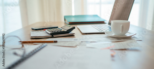 Close-up of a calculator and documents on a desk with a modern laptop in the background, next to a bright window with daylight. Business, home finances, money, and savings concept.