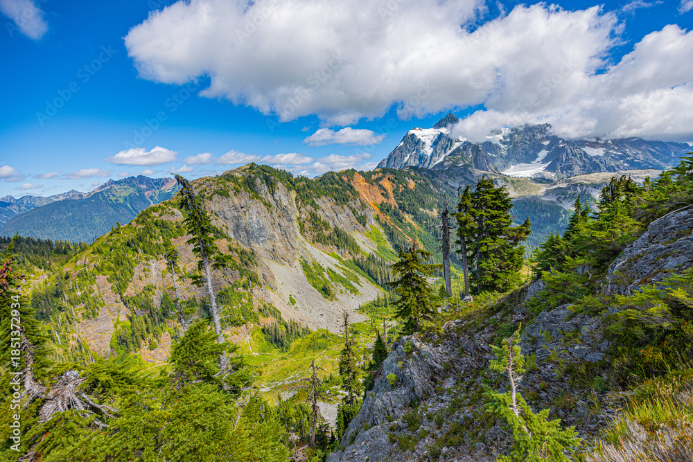 Obraz premium Rocky alpine ridge overlooking a green mountain valley with evergreen trees and distant peaks near Mount Baker in the North Cascades of Washington.