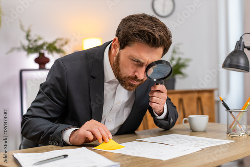 Middle-aged businessman at home office inspects contract text with magnifier finds issue and shakes head no. Freelancer guy at table writes urgent fix on sticker and places note on page to review